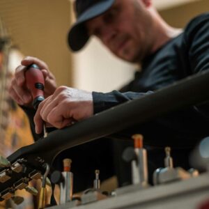 This is an image of a man in a ball cap making adjustments on the neck of an electric guitar.