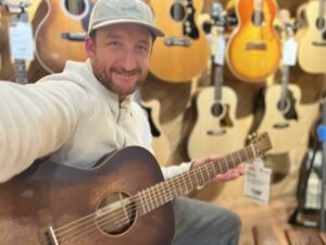 This is a photo of Brad Johnson, the writer and owner of Song Production Pros playing a Martin 000-15M StreetMaster. He is wearing a white sweatshirt and hat in a guitar shop room.