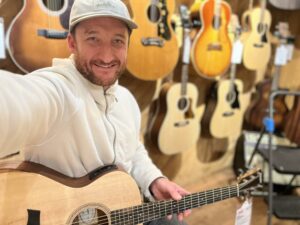 This is a photo of Brad Johnson, the writer and owner of Song Production Pros playing a Taylor Academy 12e. He is wearing a white sweatshirt and hat in a guitar shop room.