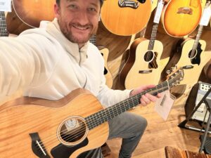 This is a photo of Brad Johnson, the writer and owner of Song Production Pros playing a Taylor GS Mini Mahogany. He is wearing a white sweatshirt and hat in a guitar shop room.