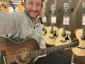 This is a photo of Brad Johnson, the writer and owner of Song Production Pros playing a Taylor 322ce. He is wearing a white sweatshirt and hat in a guitar shop room.