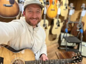 This is a photo of Brad Johnson, the writer and owner of Song Production Pros playing a Taylor 512ce Urban Red Ironbark. He is wearing a white sweatshirt and hat in a guitar shop room.