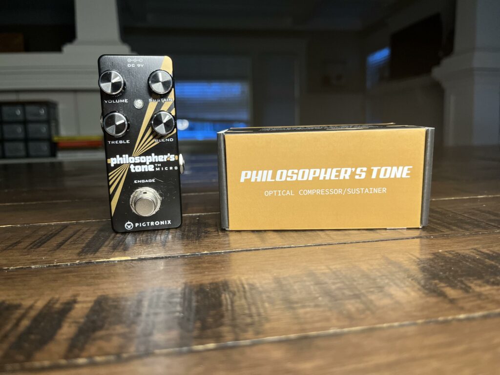 An original picture of the Pigtronix Philosopher's Tone mini guitar pedal sitting next to it's packaging on a hardwood table.