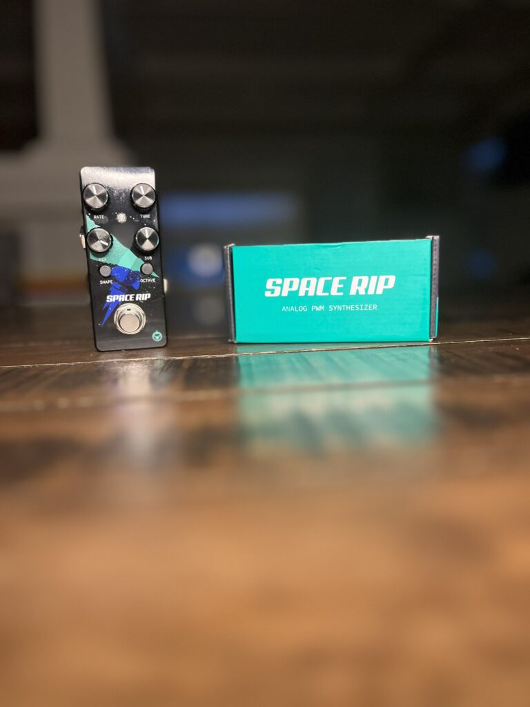 An original picture of the Pigtronix Space Rip mini guitar pedal sitting next to it's packaging on a hardwood table.