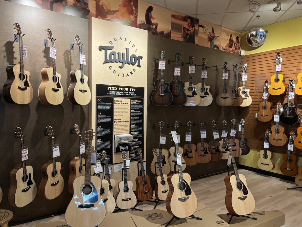 This is a photo of the Sam Ash in Westminster's Acoustic Guitar Room. There are lots of acoustic guitars lined up and a Taylor Logo on the wall.
