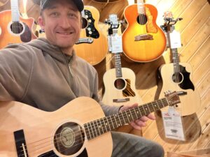 This is a photo of Brad Johnson, the writer and owner of Song Production Pros playing a Gibson G-00. He is wearing a brown sweatshirt and hat in a guitar shop room.