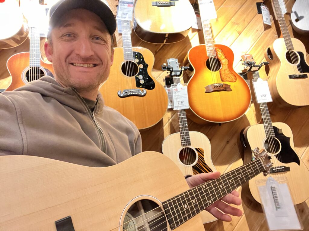 This is a photo of Brad Johnson, the writer and owner of Song Production Pros playing a Gibson G-45. He is wearing a brown sweatshirt and hat in a guitar shop room.