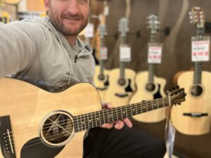 This is a photo of Brad Johnson, the writer and owner of Song Production Pros playing a Martin SC-13e.. He is wearing a grey sweatshirt and hat in a guitar shop room.