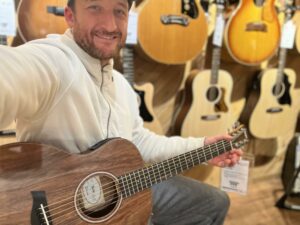 This is a photo of Brad Johnson, the writer and owner of Song Production Pros playing a Taylor GS-Mini Koa. He is wearing a white sweatshirt and hat in a guitar shop room.