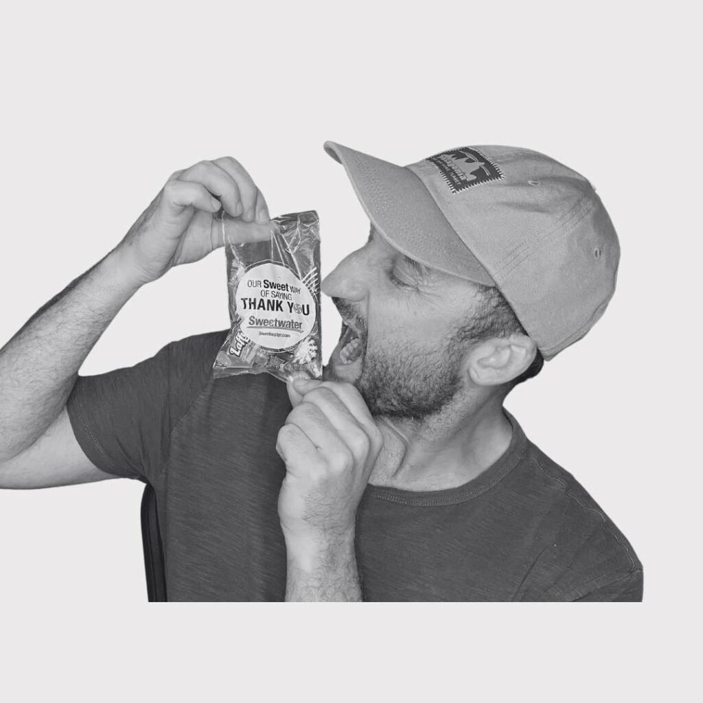 A black and white picture of Song Production Pros owner/writer biting a bag of candy that was sent by Sweetwater in a gear purchase. Brad is wearing a hat and a t-shirt.