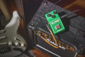 This picture shows a green Ibanez Tubescreamer guitar pedal sitting on top of a Marshall combo guitar amp. On the left side of the picture there is a silver stratocaster-style electric guitar