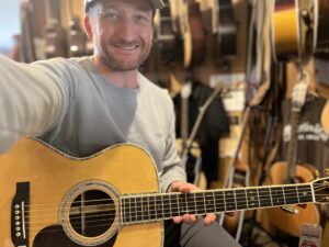 This is a photo of Brad Johnson, the writer and owner of Song Production Pros playing a Martin 000-28. He is wearing a blue sweatshirt and green hat in a guitar shop room.