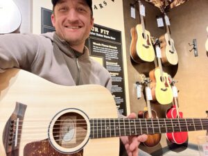 This is a photo of the founder and writer of Song Production Pros, Brad Johnson, testing out a Big Baby Taylor. This is the Solid Sitka Spruce top version. He is smiling and wearing a grey jacket and hat.
