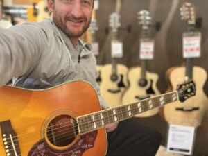 This photo shows Brad Johnson (Founder/Writer of Song Production Pros) demoing an Epiphone Hummingbird Inspired by Gibson at a guitar shop. He is wearing a grey hoody.